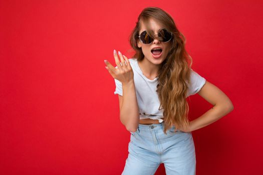 Photo shot of beautiful surprised young blonde woman wearing casual clothes and stylish sunglasses isolated over colorful background looking at camera and having questions. copy space