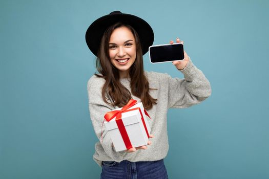 Shot of pretty smiling positive young brunette woman isolated over blue background wall wearing stylish black hat and grey sweater holding gift box showing smartphone screen display for mockup and looking at camera.