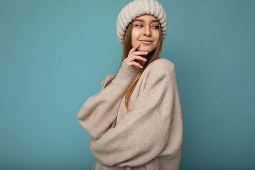 Shot of attractive happy young dark blonde woman standing isolated on blue background wall wearing beige warm sweater and winter beige hat looking to the side and having an idea. Empty space, copy space