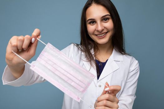 Positive smiling woman holding virus protective mask against coronavirus and wearing white medical coat isolated over blue background.