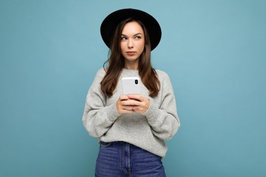 Beautiful young brunette woman thinking wearing black hat and grey sweater holding smartphone looking to the side texting isolated on background.Copy space