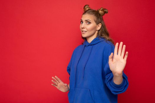 Photo of young beautiful nice blonde woman with two horns with sincere emotions wearing trendy bright blue hoodie isolated over red background with empty space and showing stop sign with hand palm.