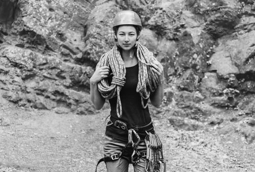 Young woman wearing in safety harness with climber equipment and helmet holding rope and looking at camera on background of rock. Monochrome image