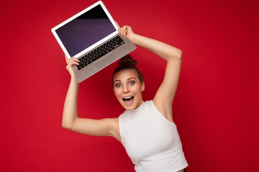 beautiful fascinating happy surorised blond young woman with gathered hair looking at camera holding computer laptop with empty monitor screen with mock up and copy space wearing white t-shirt isolated over red wall background.