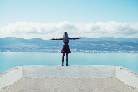 Young woman standing with raised arms on coast and looking at sea bay, rear view