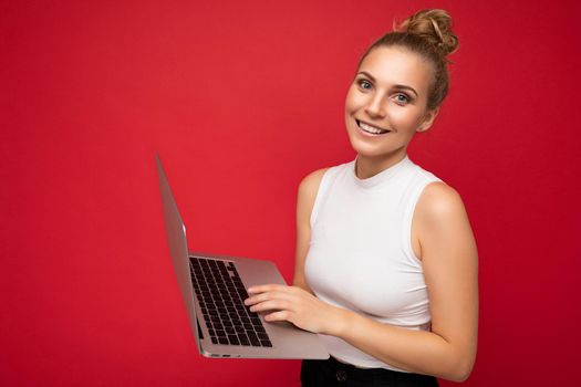 Side profile photo shot of beautiful smiling blond young woman with gathered hair wearing white t-shirt holding computer laptop typing on keyboard looking at camera isolated over red wall background.