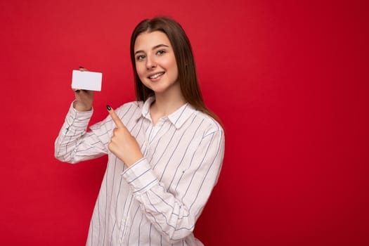 Photo shot of attractive positive smiling young dark blonde woman wearing white blouse isolated over red background holding credit card looking at camera pointing finger at plastic contactless card.