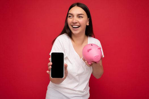 beautiful happy young brunette woman wearing casual white t-shirt isolated over red background with empty space holding in hand mobile phone and showing smartphone with empty screen for mockup and money box looking like pink pig looking to the side.
