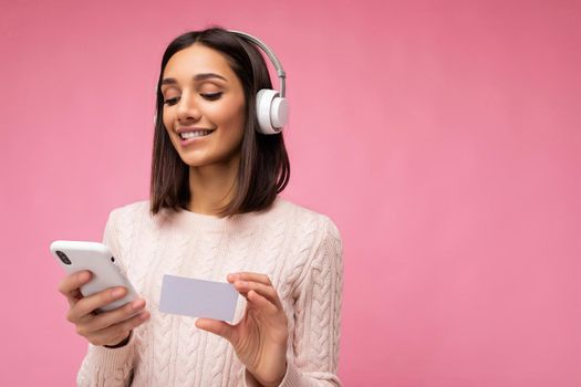 Photo of beautiful positive smiling young brunette woman wearing pink casual sweater isolated over pink background wall wearing white bluetooth wireless headphones and listening to music and using mobile phone making payment online through credit card looking down. copy space