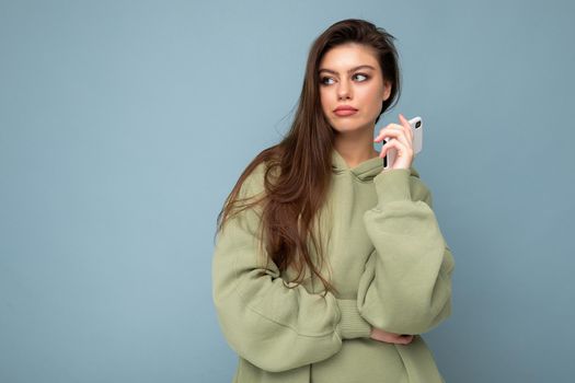 Concentrated upset attractive young woman wearing stylish hoodie holding using mobile phone isolated on background looking to the side having an idea and dreaming. Copy space