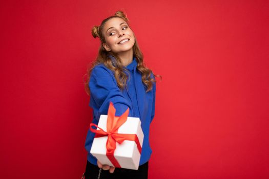 Fascinating happy smiling amazing young blonde woman isolated over red background wall wearing blue stylish hoodie holding gift box and looking at camera.
