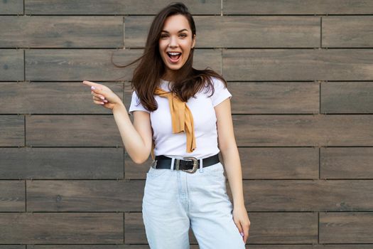 Young emotional positive joyful smiling beautiful brunette woman wearing casual clothes standing in the street near the wall and pointing at empty space. Lifestyle concept.