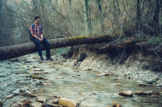 Traveler young man sitting on tree trunk over river in autumn forest