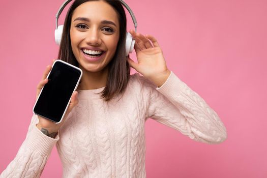 Closeup portrait of Beautiful cute young brunette woman wearing pink sweater isolated over pink background wall wearing white bluetooth wireless headphones and listening to good music and showing mobile phone with empty display for mockup having fun.