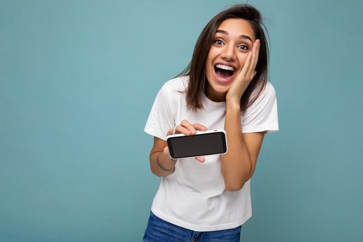 Photo of pretty positive shocked young brunette woman good looking wearing white t-shirt standing isolated on blue background with copy space holding smartphone showing phone in hand with empty screen screen for cutout.