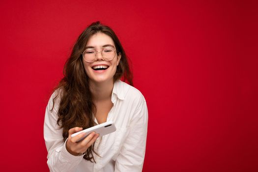photo of Pretty smiling positive young brunette woman wearing white shirt and optical glasses isolated over red background holding in hand and using mobile phone playing online games looking at camera and having fun.