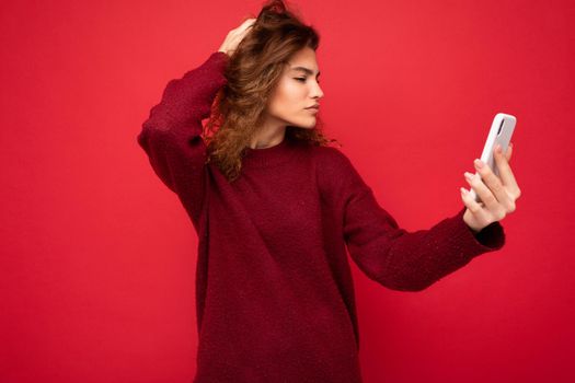 Attractive young blogger woman with curly hair wearing dark red sweater isolated on red background wall holding and using smart phone looking at telephone screen and taking selfie.