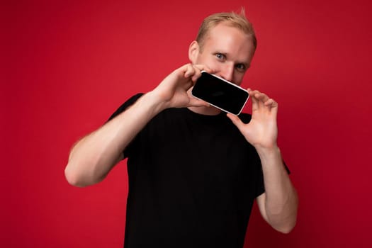 Handsome happy cool young man good looking wearing casual stylish clothes standing isolated over colourful background wall holding smartphone and showing phone with empty screen display looking at camera.
