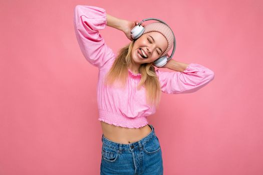 Photo of attractive positive smiling young blonde woman wearing pink blouse and pink hat isolated over pink background wall wearing white wireless bluetooth earphones listening to music having fun.