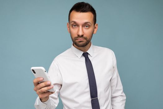Closeup portrait of handsome good looking brunet unshaven man with beard wearing casual white shirt and tie isolated on blue background with empty space holding in hand mobile phone communicating via smartphone on the internet looking at camera.