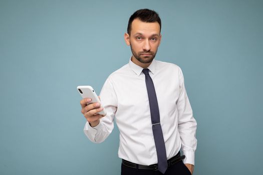 Handsome good looking brunet unshaven man with beard wearing casual white shirt and tie isolated on blue background with empty space holding in hand mobile phone communicating via smartphone on the internet looking at camera.