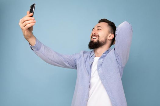 Self-confident handsome young brunet bearded man wearing casual stylish clothes standing isolated over blue background wall holding smartphone taking selfie photo looking at mobile phone screen display.