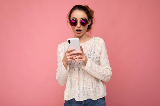 Photo of attractive dissatisfied shocked young woman wearing casual white blouse and colorful sunglasses isolated over pink background wall holding and using phone looking at gadjet display.