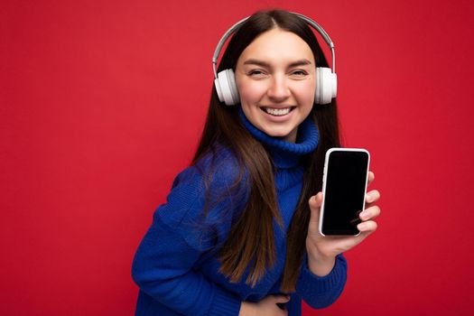 Beautiful happy smiling young woman wearing stylish casual outfit isolated on background wall holding and showing mobile phone with empty display for mockup wearing white bluetooth headphones listening to music and having fun looking at camera.