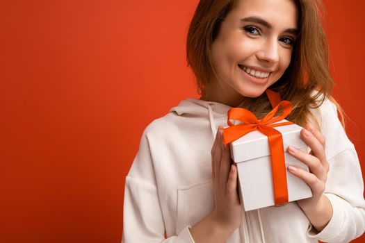 Closeup photo shot of acharming sexy positive smiling blonde young woman isolated over colorful background wall wearing white hoodie holding white gift box with red ribbon and looking at camera.