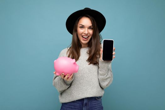 Portrait photo of happy positive smiling satisfied sincere young attractive brunette woman wearing stylish grey sweater and black hat isolated on blue background with free space and holding pink pig penny bank and showing mobile phone with empty screen display.