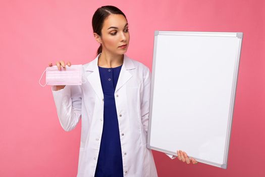 Woman doctor in a white medical coat holding blank board with copy space for text and protective mask isolated on background. Covid concept.