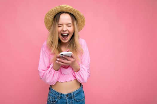 Attractive amazed young blonde woman smiling and having fun standing isolated over pink wall keeping hand in pocket wearing pink blouse and summer hat using mobile phone with close eyes.