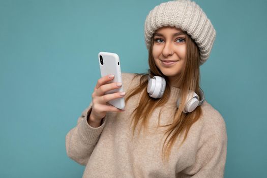 Closeup Photo of beautiful happy smiling young dark blonde woman wearing beige stylish sweater and knitted beige winter hat isolated on blue background wearing white bluetooth wireless headphones and listening to cool music and using mobile phone surfing on the internet looking at camera.