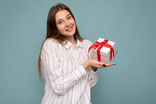 Beautiful happy young dark blonde woman isolated over colourful background wall wearing stylish casual clothes holding gift box and looking at camera.