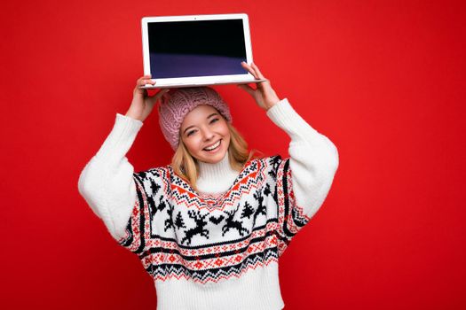 Close-up portrait of Beautiful young woman holding computer laptop with empty monitor screen with mock up wearing knitted winter hat and sweater looking at camera isolated over red wall background.