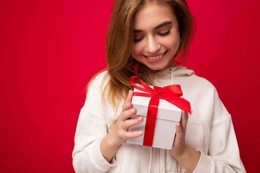 Photo shot of pretty positive smiling young dark blond woman isolated over colourful background wall wearing trendy outfit look holding gift box and looking at present box with red ribbon.