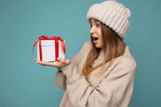 Photo shot of pretty positive surprised young dark blond woman isolated over colourful background wall wearing trendy outfit look holding gift box and looking at present box with red ribbon.