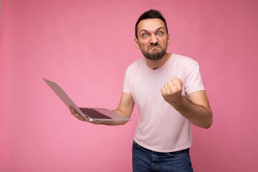 Handsome angry and wrathful man holding laptop computer showing fist looking at camera in t-shirt on isolated pink background.