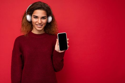 Beautiful happy smiling young woman wearing stylish casual outfit isolated on background wall holding and showing mobile phone with empty display for mockup wearing white bluetooth headphones listening to music and having fun looking at camera.