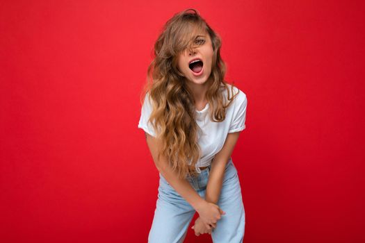 Shot of young emotional happy joyful pretty blonde woman wearing white t-shirt isolated on red background with empty space and shouting.