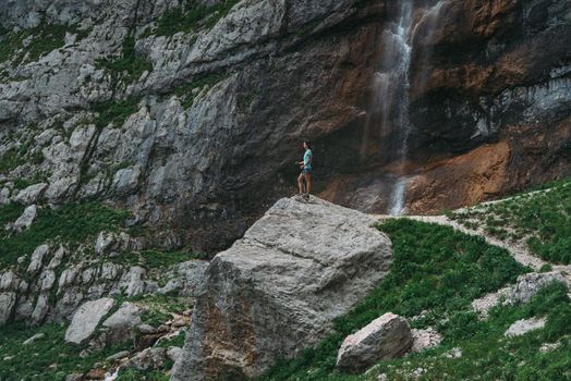 Traveler young woman with trekking poles standing on stone on background of rocks and waterfall