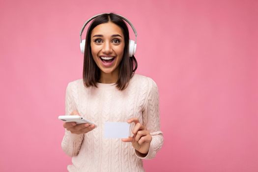 Photo of beautiful young smiling brunette woman wearing pink casual sweater isolated over pink background wall wearing white bluetooth wireless headphones and listening to music and using mobile phone making payment online through credit card looking at camera and having fun.