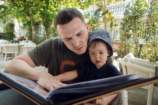 Adult man with adorable toddler sitting at desk and watching book on terrace of hotel with green plants on background.