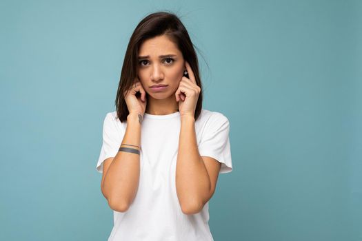 Portrait of sad sorrowful young pretty nice brunette woman with sincere emotions wearing casual white t-shirt for mockup isolated on blue background with copy space and covering ears with fingers.
