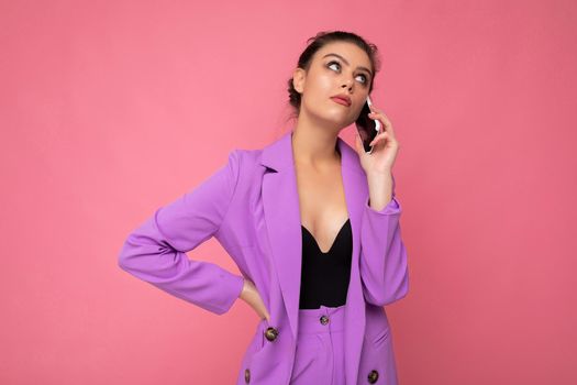Photo of sexy beautiful young woman wearing purple suit talking on the mobile phone isolated over pink background looking up and rolling eyes.