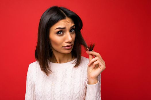 Portrait of attractive sad young brunette woman wearing sweater isolated on red background with copy space and touching short hair and having problems with split ends.