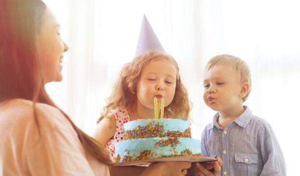 Cheerful woman holding festive cake and siblings blowing candles in backlit