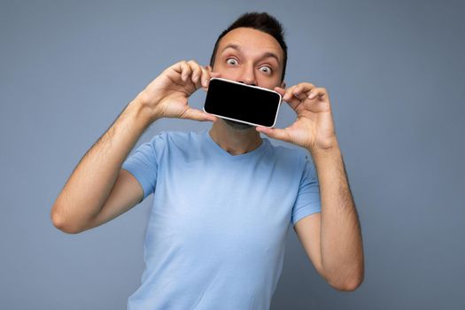 Handsome happy cool young man good looking wearing casual stylish clothes standing isolated over colourful background wall holding smartphone and showing phone with empty screen display looking at camera.