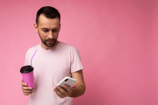 Photo shot of handsome positive good looking young man wearing casual stylish outfit poising isolated on background with empty space holding in hand and using mobile phone messaging sms looking at smartphone display screen.