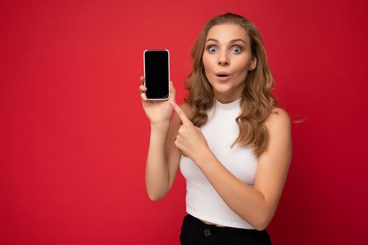 Amazed surprised beautiful young blonde woman wearing white t-shirt isolated on red background with copy space holding smartphone showing phone in hand with empty screen for mockup looking at camera and pointing at gadjet.